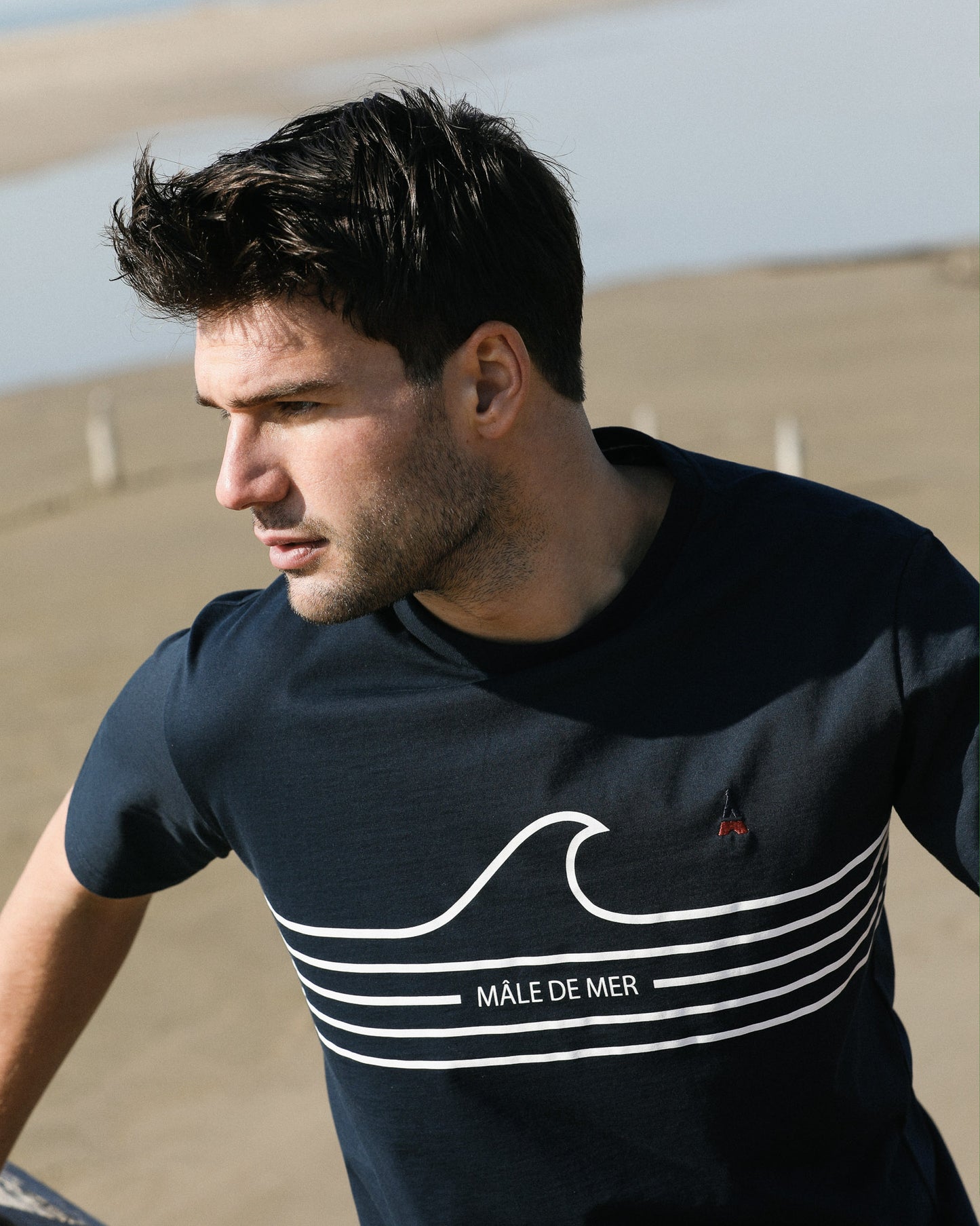 Homme debout sur la plage portant un tee-shirt bio éco-responsable bleu marine écrit mâle de mer avec une vague